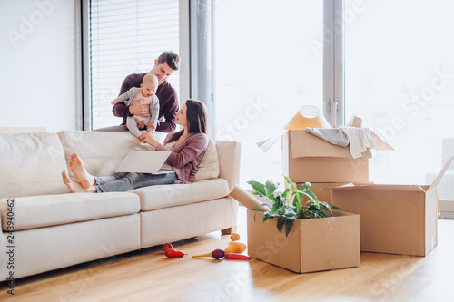 A portrait of young couple with a baby and cardboard boxes moving in a new home фототапет