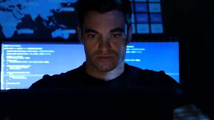 close up portrait of developer programmer looks at camera in a data center filled with monitor screens. Monitor screen is reflected on his face