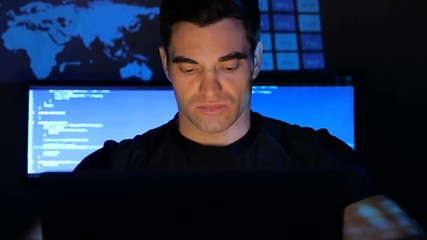 close up portrait of developer programmer looks at camera in a data center filled with monitor screens. Monitor screen is reflected on his face