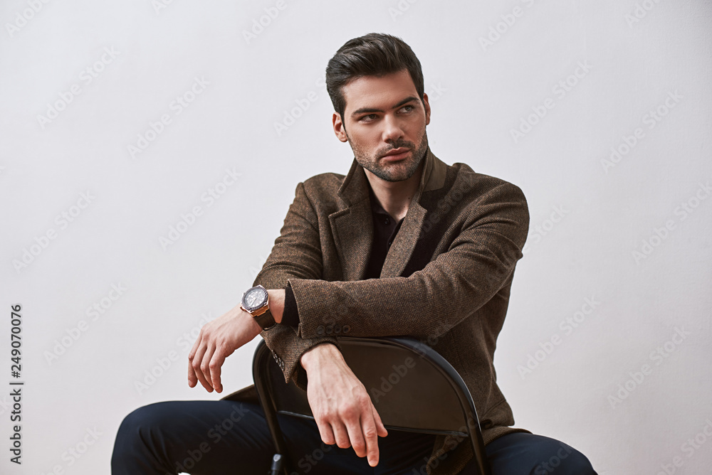 Perfect style. Stylish dark-haired man sitting on a chair and looking away isolated over white studio background - obrazy, fototapety, plakaty 