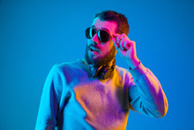 Enjoying His Favorite Music. Happy Young Stylish Man In Hat And Sunglasses With Headphones Listening And Smiling While Standing Against Blue Neon Background