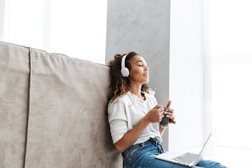 Wall Mural - Image of attractive african american girl wearing headphones drinking tea and using laptop, while sitting on floor in bright living room