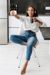Wall Mural - Image of attractive african american girl wearing headphones taking selfie photo, while sitting in bright kitchen