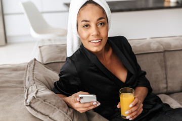 Wall Mural - Portrait of gorgeous african american woman wearing housecoat, using mobile phone and drinking juice at home