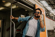 © KleverLevel - Attractive smiling young hispanic wearing sunglasses and blue shirt male holding smartphone hand and calling to friends while he standing at the train wagon on platform.