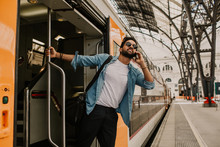 Confident Attractive Young Hispanic Wearing Sunglasses And Blue Shirt Male Holding Smartphone Hand And Calling To Friends While He Standing At The Train Wagon On Platform.