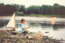 Blond Boy  Fishing On The Lake. He Is Sitting On A Raft With A Sail In Summer Day.