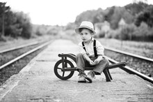 Monochrome Portrait Of Cute Boy Wearing In Retro Style Sitting On Railway Station And Is Waiting For Something. 