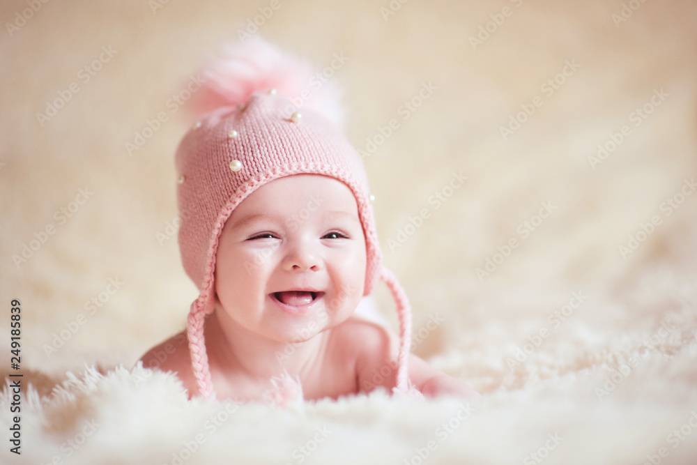 Happy baby girl wearing stylish knitted hat lying in bed close up. Childhood. - obrazy, fototapety, plakaty 