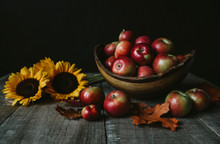 Close-up Of Fresh Apples With Sunflowers And Leaves On Wooden Table Against Black Background