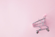 Small Supermarket Grocery Push Cart For Shopping Toy With Wheels And Pink Plastic Elements On Pink Pastel Color Paper Flat Lay Background. Concept Of Shopping. Copy Space For Advertisement