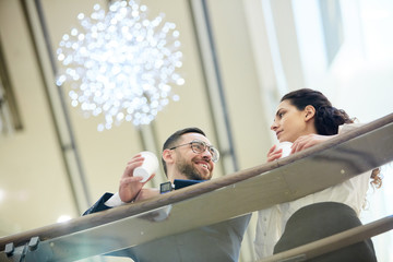 Wall Mural - Young successful businessman and his colleague with drinks standing by railings inside mall and talking