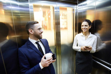 Wall Mural - Happy millennials in formalwear looking at each other while using smartphones inside elevator