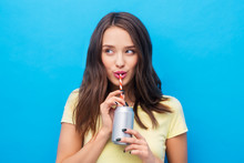 People Concept - Smiling Young Woman Or Teenage Girl In Yellow T-shirt Drinking Soda From Can Through Paper Straw Over Bright Blue Background