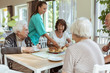 © Maskot - Young nurse serving food and drink to elderly people at table in nursing home