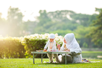 Muslim mother teaching her little son to drawing and painting with crayons in the garden on grass field . Muslim family concept.