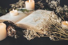 Close Up Of Open Vintage Poetry Book Decorated With Dried Baby's Breath Flowers. Blurred Background With White Lit Burning Candles, Plants And Small Sage Stick. Black Table Surface. Romantic Soft Feel