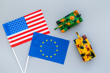 War, Confrontation Concept. European Union, USA. Tanks Toy Near European And American Flag On Grey Background Top View