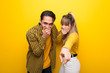 © luismolinero - Young couple over vibrant yellow background pointing with finger at someone and laughing