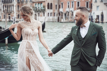 Happy wedding couple near the canal in Venice. Elegant woman in luxury ivory dress, messy updo hair, man in green three-piece suit and bow tie