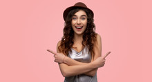 Indoor Shot Of Attractive Girl Crosses Arms Over Chest, Indicates At Different Sides With Fore Fingers, Hesitates Between Two Items In Shop, Isolated On Pink Wall 