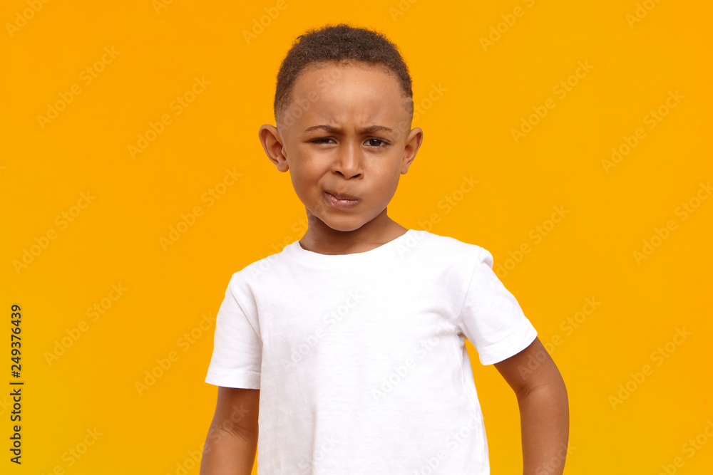 Studio image of grumpy dissatisfied dark skinned black little boy in white t-shirt frowning, being dissatisfied with unfair grade at school, looking at camera with confused facial expression - obrazy, fototapety, plakaty 