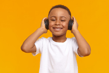 Wall Mural - Technology, leisure, music and entertainment concept. Picture of adorable cute African child with broad smile, enjoying favorite songs using wireless headphones. Black little boy listening to music