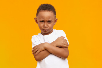 Wall Mural - Isolated image of funny cool African American little boy frowning and keeping arms crossed on his chest against blank yellow studio wall with copy space for your text. Life perception and attituve