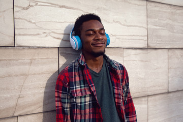 Wall Mural - Portrait urban smiling african man in wireless headphones enjoying listening to music on gray wall background, wearing plaid shirt