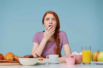 Wall Mural - Frightened scared red-haired girl covers open mouth with a palm, sits at a table on which there are many different foods baking fresh fruit,worrying about her health figure, diet violation