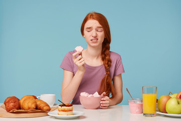 Wall Mural - A red-haired girl sitting at a table during breakfast tried a fruit marshmallow, holding a piece in her hands looks at him with disgust, she didn’t like the taste isolated on a blue background.