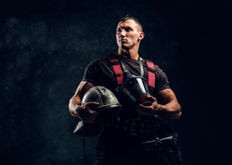 Wall Mural - Portrait of a handsome muscular fireman holding a helmet and oxygen mask standing in the studio against a dark textured wall