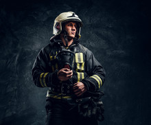 Studio Portrait In A Dark Studio Against A Textured Wall. Brutal Firefighter In Uniform And Safety Helmet Holding An Oxygen Mask And Looking Sideways With A Confident Look.