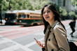 © PR Image Factory - beautiful college girl smiling looking aside holding cellphone while crossing road on street in san francisco. bus light rail train in background. happy young woman having fun chatting on smart phone