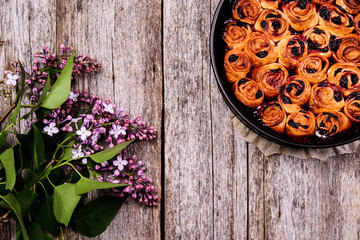 Wall Mural - Homemade mini cherry rolls in baking form with bouquet of lilac on vintage wooden table. Bun cake with cherry filling