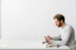 © Joske - A thirty something man with a red beard and curly hair in a grey pull over is looking on his smart phone while working on his laptop in a clean white empty office setting.