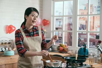 happy asian woman housewife in apron cooking making chocolate melting with spoon in pot on stove in wooden kitchen decorated with heart balloons. young girl handmade sweet dessert  for valentine day.