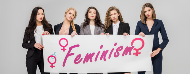 Wall Mural - attractive businesswomen holding  large sign with feminism lettering isolated on grey