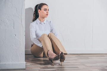 Wall Mural - upset woman in white blouse and beige pants sitting on floor near white wall at home, grieving disorder concept