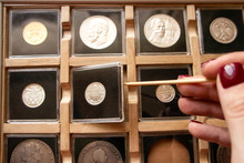 Female Hand Taking Out A Coin From A Wooden Display Case With Numismatic Collection With A Wooden Stick. Coin Holder Case With Plastic Protection Square Capsules