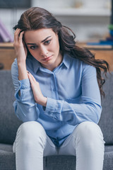 Poster - sad woman in blue blouse and white pants sitting on grey couch at home, grieving disorder concept