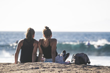 People Sitting Seafront Looking The Ocean. Two Young Girls Relaxing On A Sit Overlooking The Sea.  Couple Of Women Enjoying The View. Back Side Of Friends Relaxing Outdoors Looking Waves And Surfers