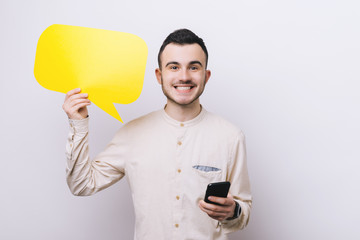 Wall Mural - Young man standing isolated over white background holding speech bubble using phone while looking camera