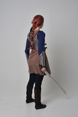 Poster - full length portrait of a  red haired girl wearing medieval warrior costume and steel armour, standing pose facing away from the camera on grey studio background.