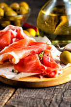 Closeup Of Thin Slices Of Prosciutto With Mixed Olives And Paprika On Wooden Cutting Board