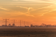 Early Morning Hazy Rural Landscape With The Silhouettes Of Transmission Towers, Trees And Farm Houses On The Background And A Bright Orange And Yellow Sky