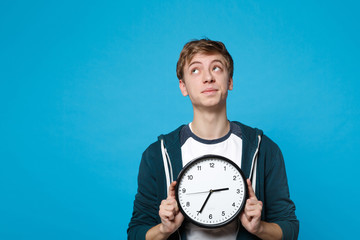 Portrait of pensive young man in casual clothes looking up, holding round clock isolated on blue wall background. Time is running out. People sincere emotions, lifestyle concept. Mock up copy space.