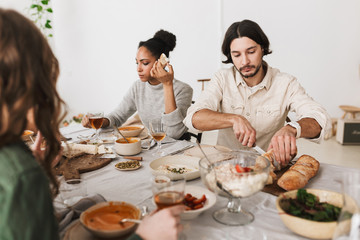 Wall Mural - Thoughtful man with dark hair and beard sitting at the table cutting bread while afro american woman near dreamily eating. Group of young international friends together on lunch in cozy cafe