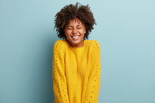 Black Overemotive Young Woman With Curly Hair, Smiles Broadly, Closes Eyes, Dressed In Yellow Sweater, Isolated Over Blue Background, Laughs At Something Pleasant, Feels Energetic And Pleased