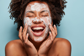 Canvas Print - Portrait of beautiful woman with white foam mask on face, touches cheeks, enjoys softness of skin after beauty treatments, has curly hair, shows bare shoulders, isolated over blue background